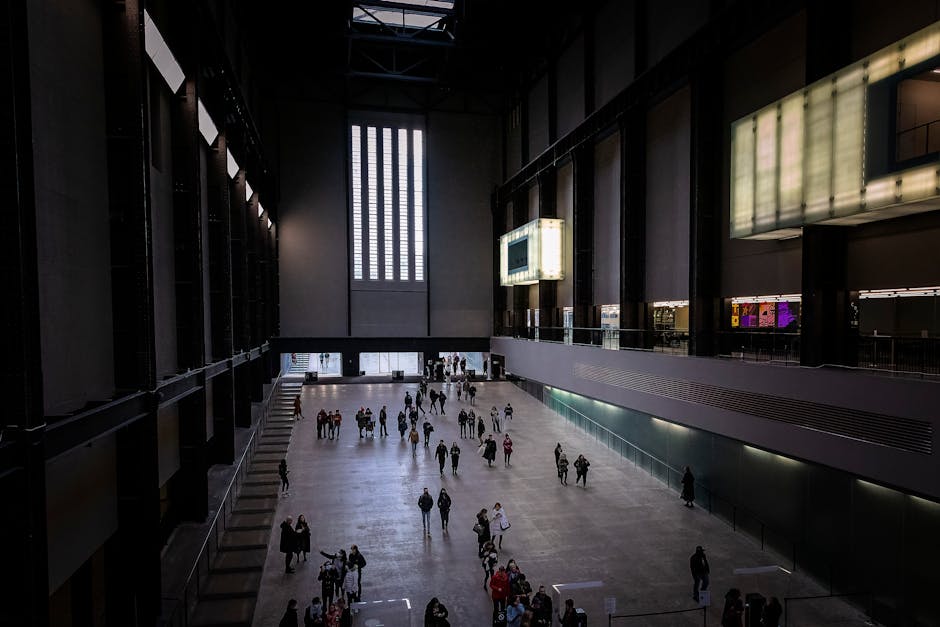 Interior view of the Tate Britain exhibition hall showing a spacious, clean floor made of smooth, polished concrete with a few scattered visitors walking and standing. The high ceiling features large, vertical windows allowing natural light to illuminate the space. The walls are dark with modern design elements, including digital display screens and large artwork frames. The area appears well-maintained with no visible dust or dirt, reflecting thorough surface cleaning and sanitisation, consistent with professional cleaning standards by Pimlico Cleaners for gallery and event prep.