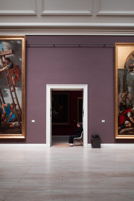 The image depicts a gallery room with a deep purple wall, featuring two large, framed classical paintings on either side. The flooring is light-colored wood, appearing clean and well-maintained. In the center, a doorway with a white frame leads to another room where a person is seated on a chair, looking at their phone or reading. To the right of the doorway, there is a small black trolley with cleaning supplies, indicating recent or ongoing cleaning activity. The lighting in the room is bright, highlighting the sheen and cleanliness of the polished wooden floors and the matte finish of the painted wall. No dust or clutter is visible, suggesting careful surface cleaning and preservation suitable for a high-value art space. Pimlico Cleaners, a professional cleaning company, offers deep cleaning and surface sanitisation services that ensure galleries like this are meticulously maintained in preparation for events and exhibitions, aligning with Tate Britain cleaning needs for event and gallery prep.
